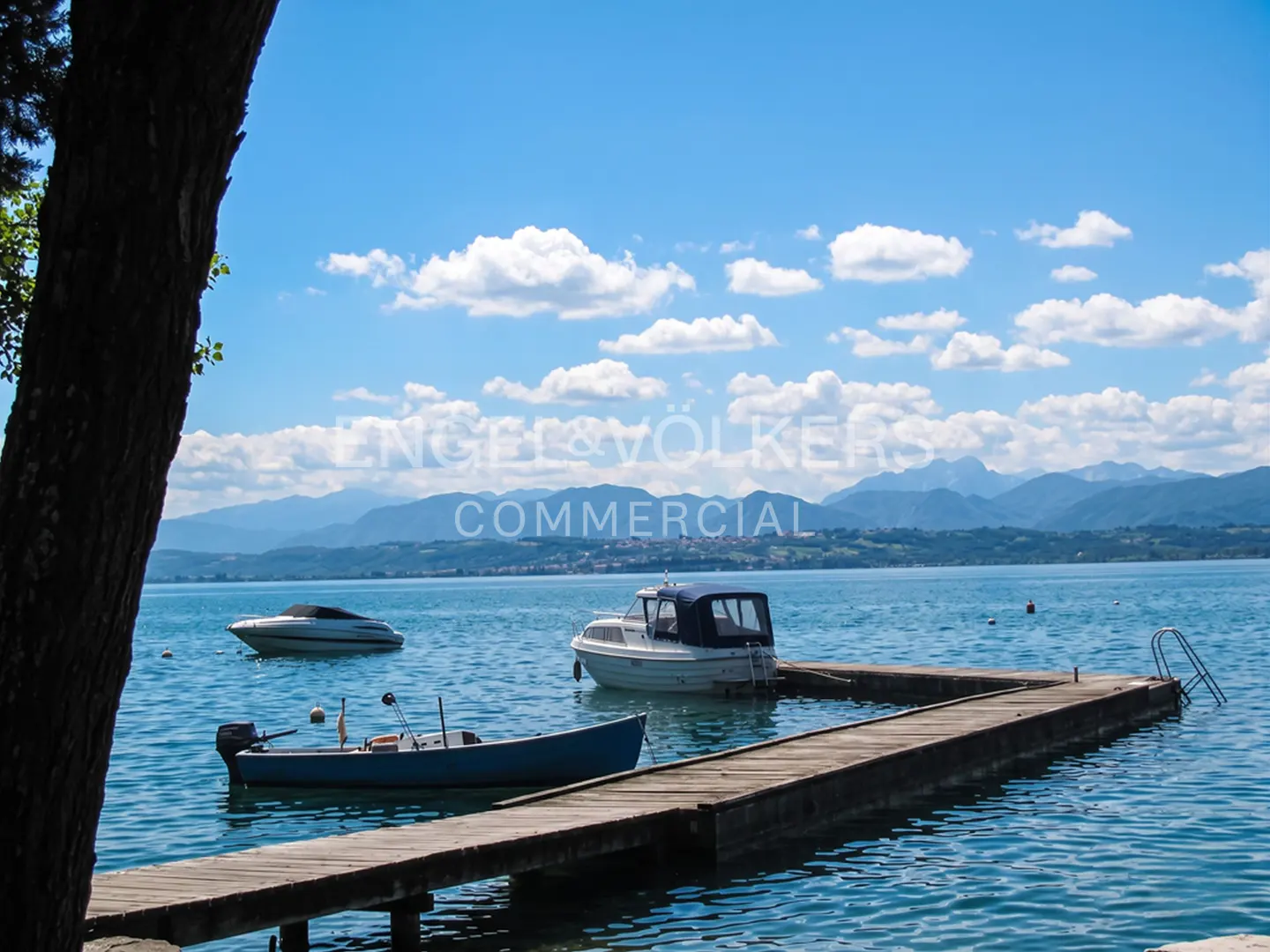 Scenic view of a lake with boats docked at a wooden pier under a blue sky with scattered clouds. Mountains are visible in the distance.