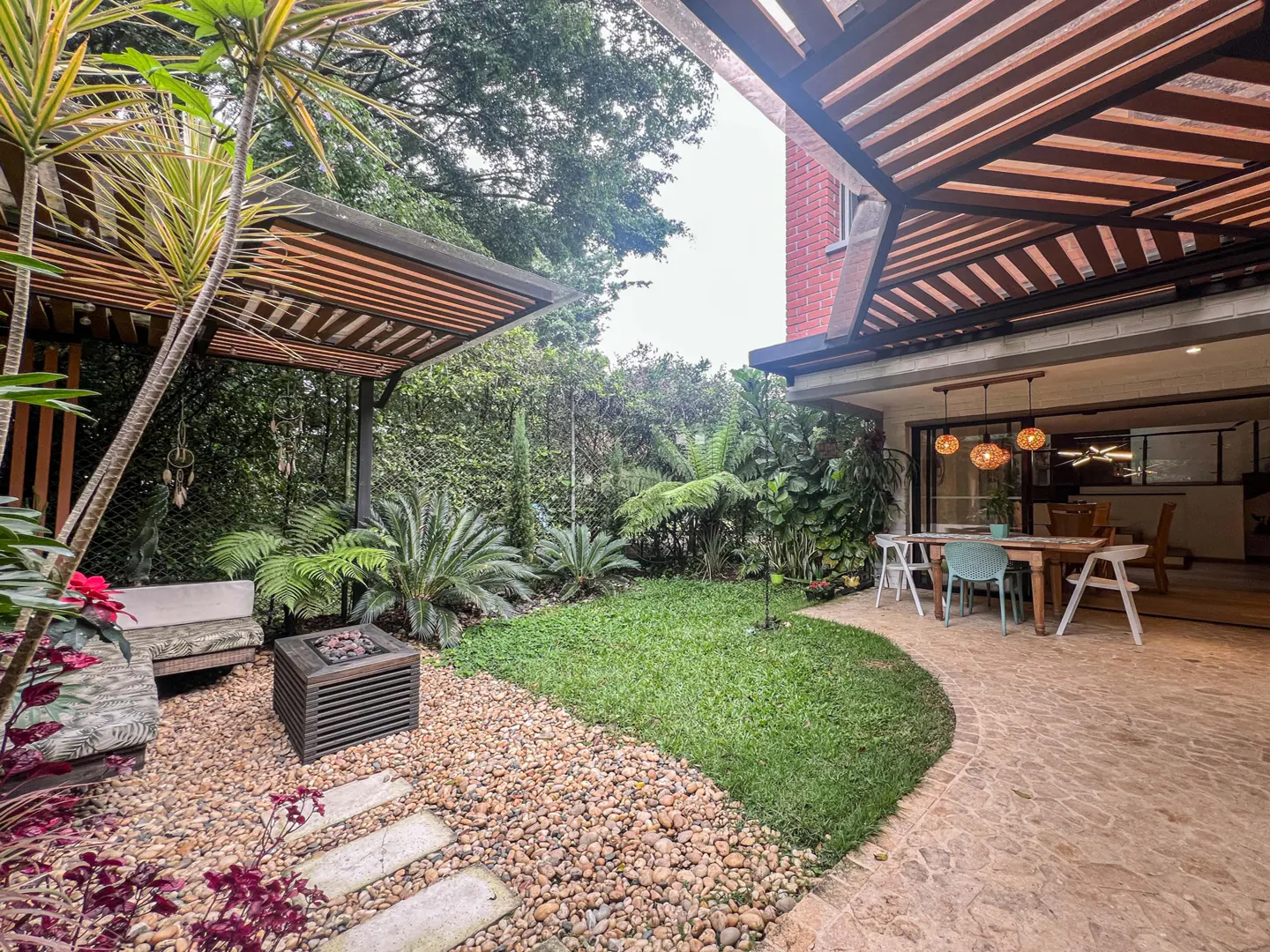 Outdoor patio with a stone fire pit, bench, and dining table under a wood pergola, surrounded by lush green plants.