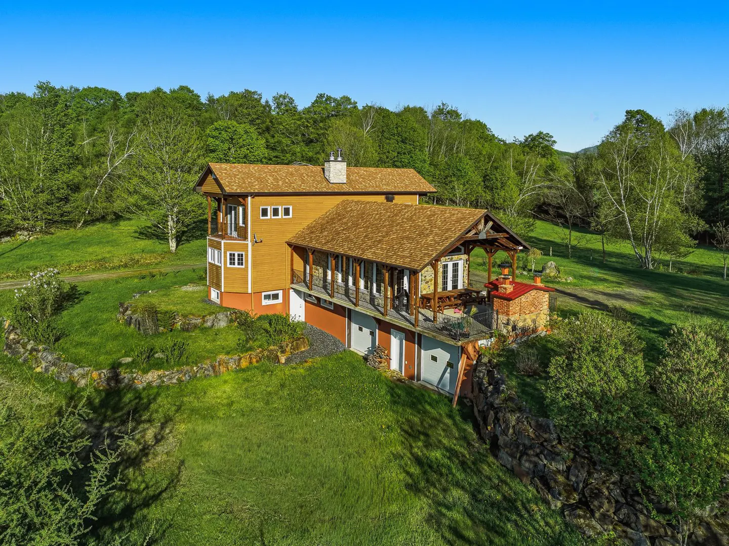 Two-story house with a brown roof and yellow siding on a green hillside with trees and a blue sky.
