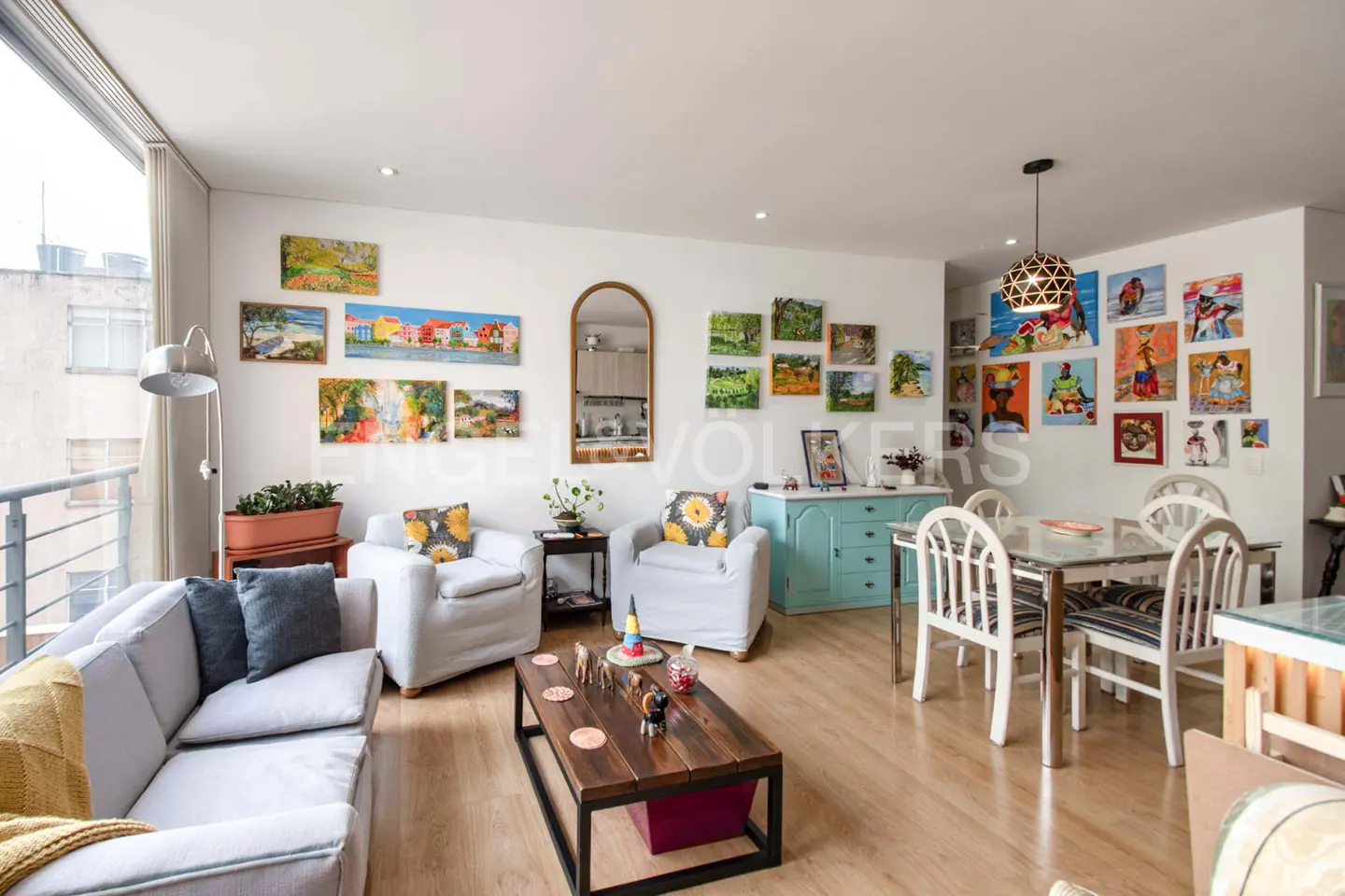 Bright living room with wood floors, white walls covered in art, a gray sofa, and a dining table with white chairs.