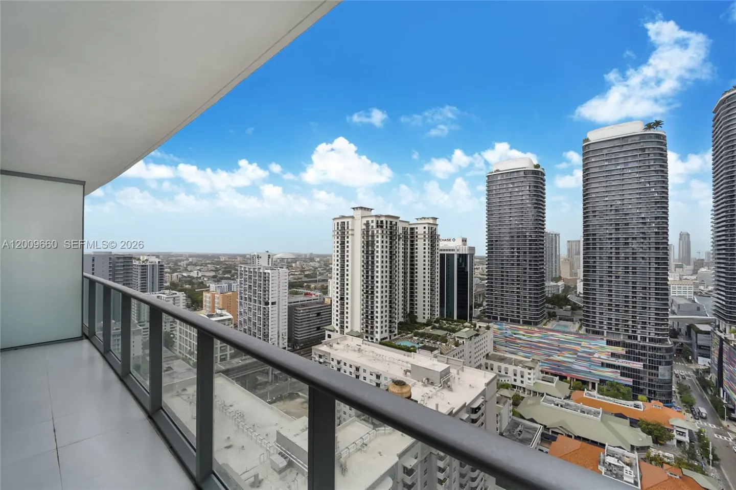 View from a high-rise balcony with glass railings overlooking a cityscape under a blue sky with scattered clouds.