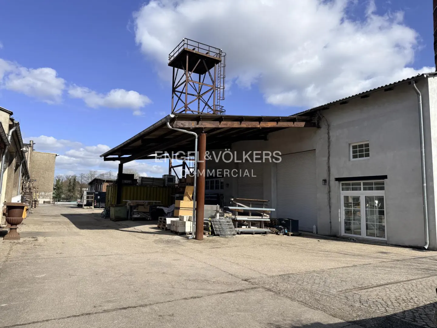 Commercial property with a metal tower on the roof, a loading dock, and a large paved yard under a blue sky with clouds.