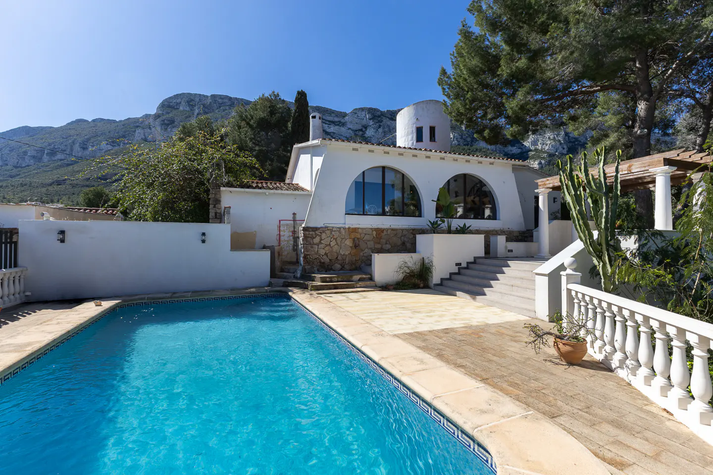 A bright, sunny view of a white villa with a blue swimming pool and mountain backdrop.