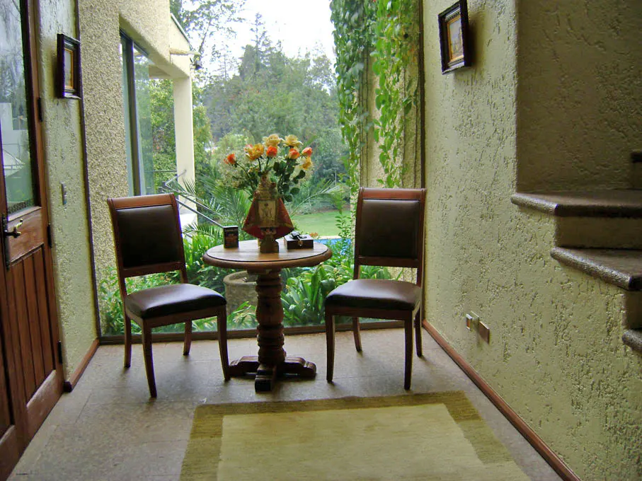 A sunroom with two brown chairs and a round wooden table with flowers. A large window overlooks a green garden.