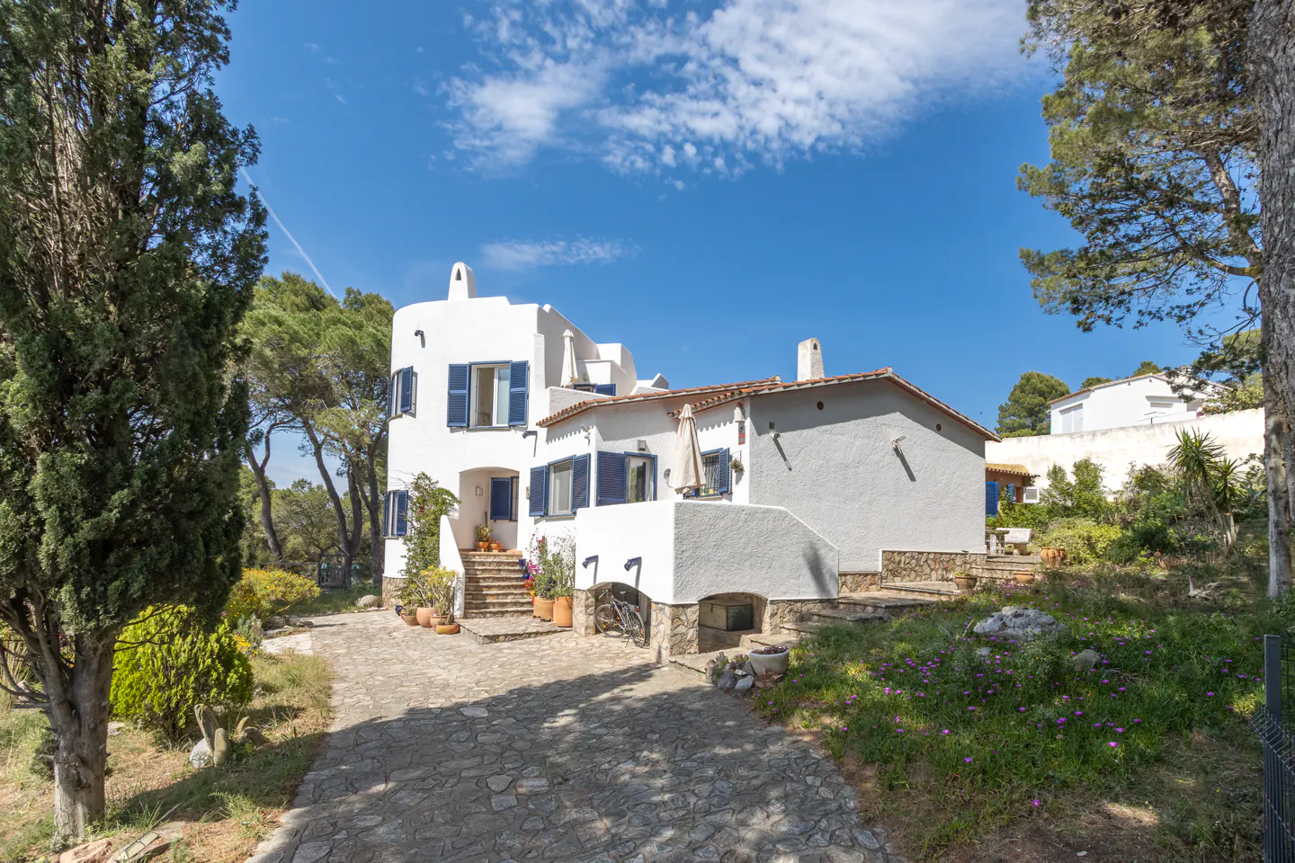 Exterior of a white house with blue shutters, surrounded by trees and a stone driveway.