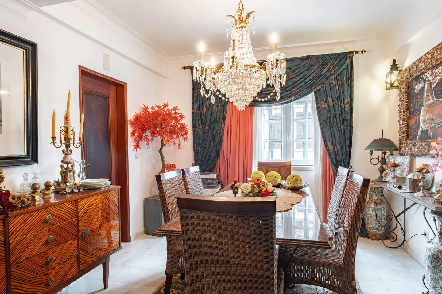 A dining room with a glass table, wicker chairs, and a crystal chandelier. A wooden sideboard and console table flank the room.