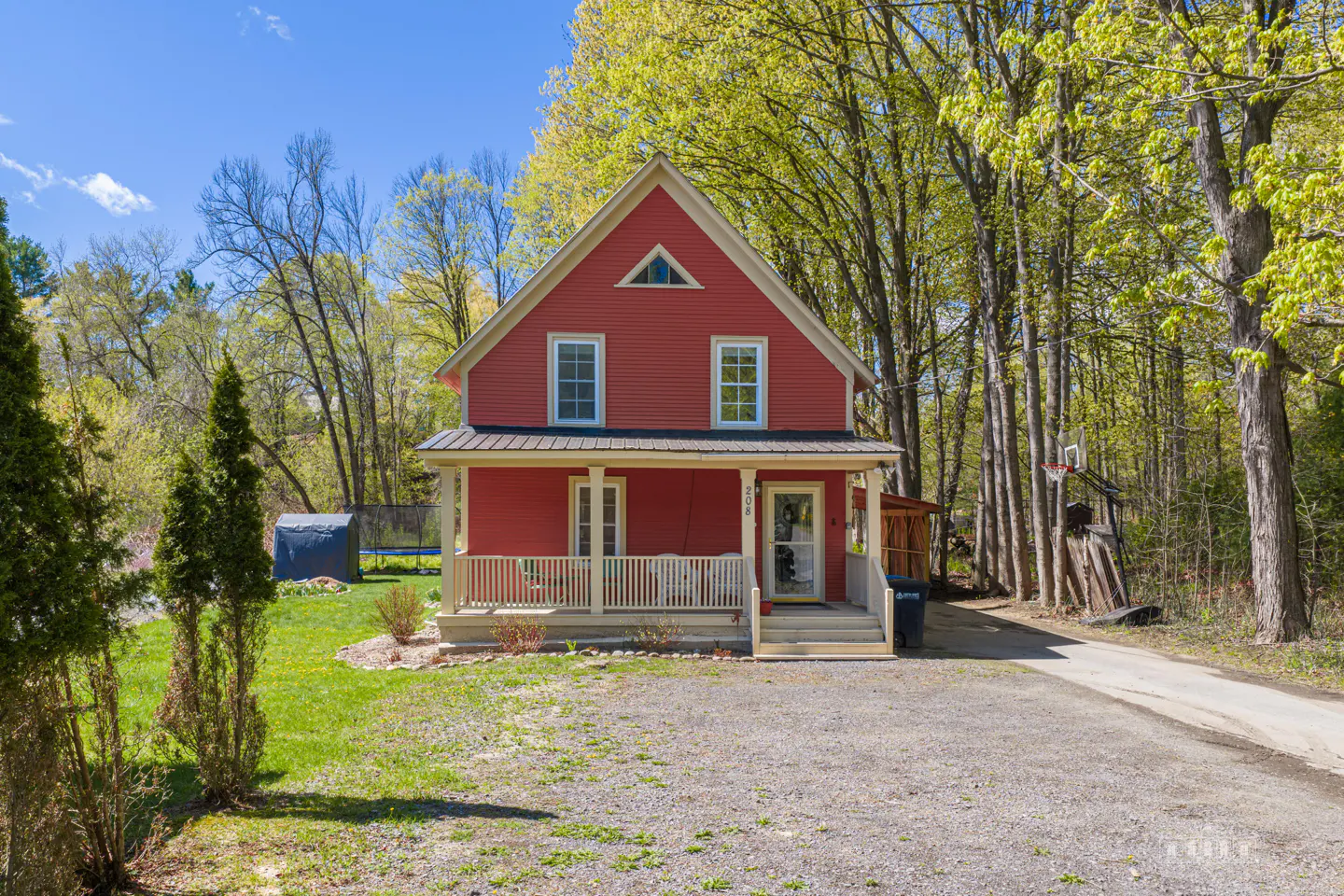 Two-story red house with a white porch and trim, surrounded by green trees and a gravel driveway.