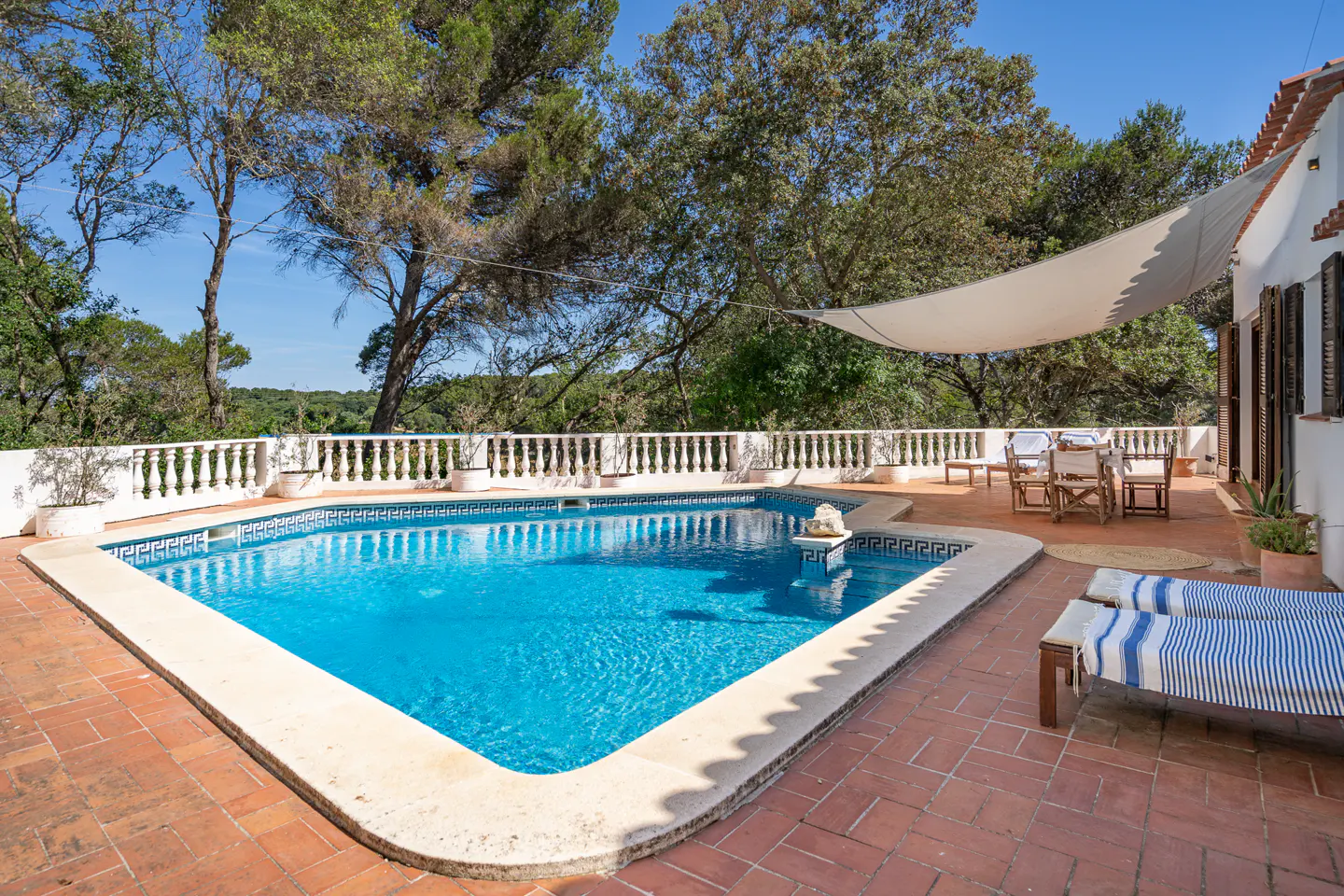 Outdoor pool with blue water, surrounded by a brick patio, white balustrade, trees, and a white sunshade.