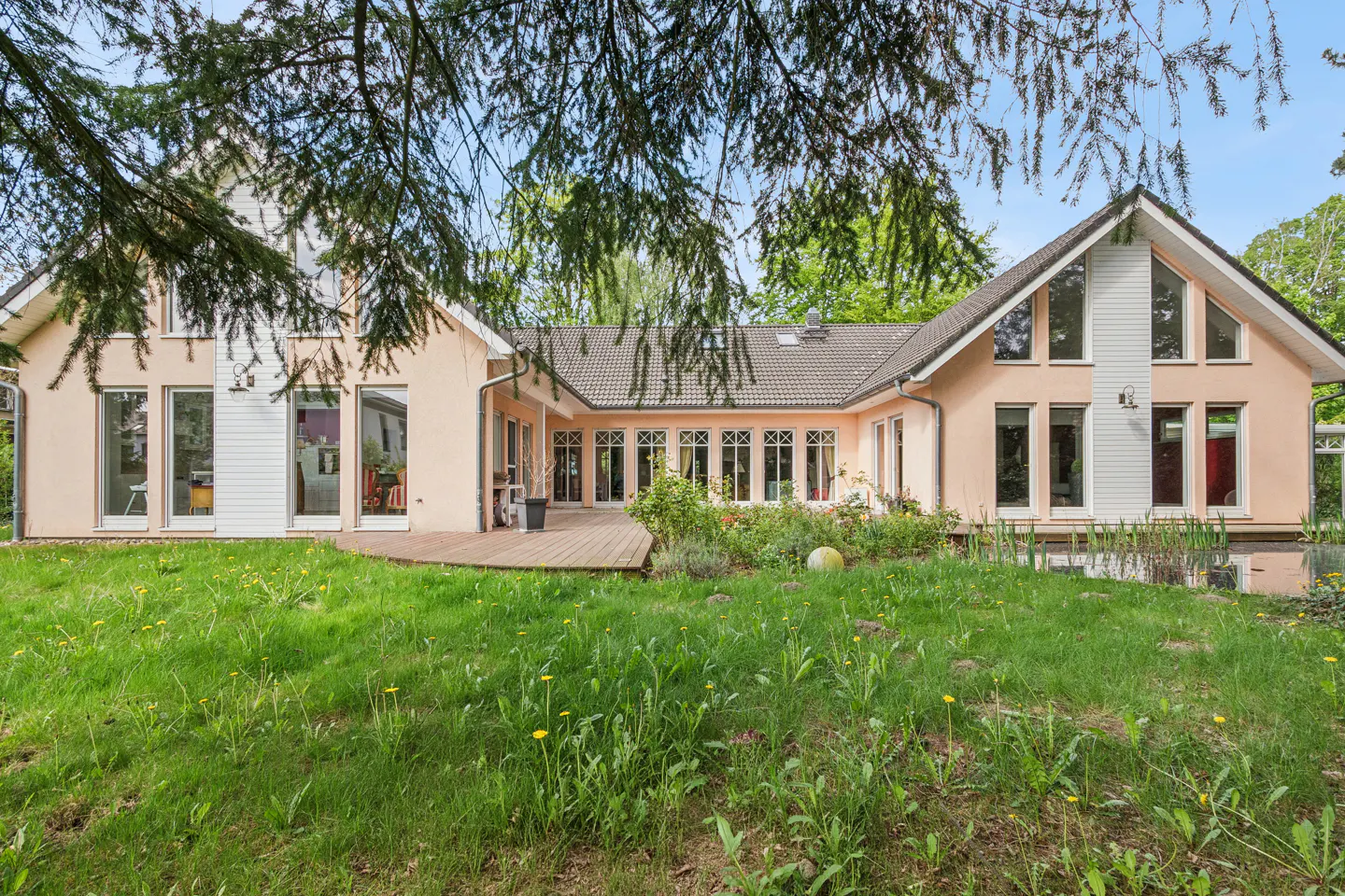 A peach-colored house with large windows and a wooden deck, set behind a grassy lawn with yellow wildflowers.