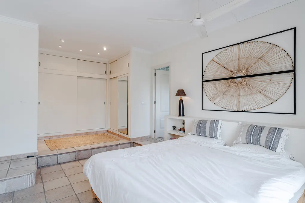 Bright bedroom with white walls, a white bed with striped pillows, and a large circular wall decoration. A closet with sliding doors is visible in the background.