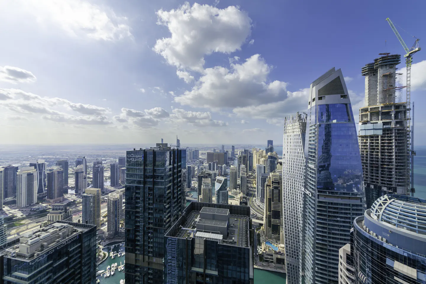 Cityscape view of Dubai with modern skyscrapers under a blue sky with white clouds. A building is under construction with a crane.