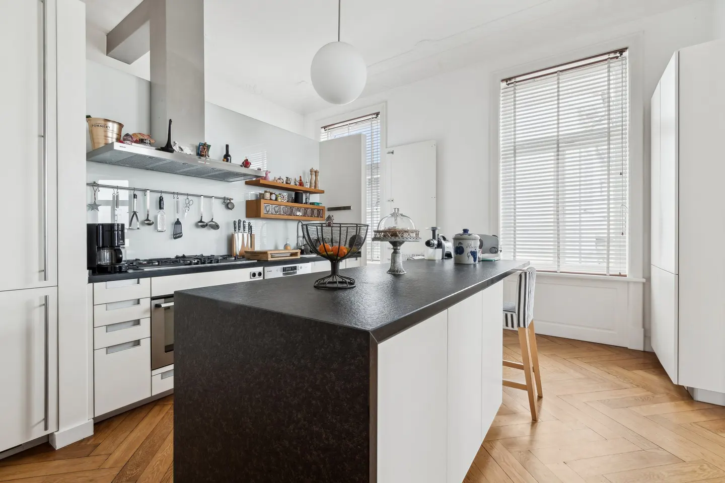 Bright kitchen with white cabinets, black countertop island, and herringbone wood floors. Utensils hang above the stove.
