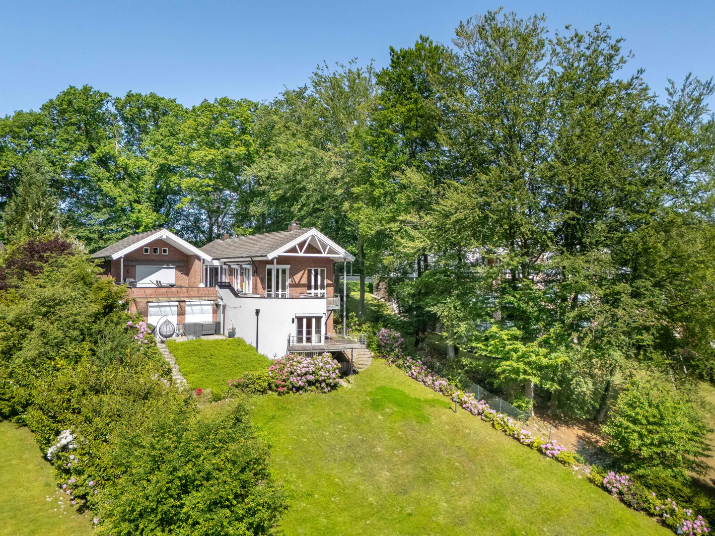 A two-story brick and white house with a large green lawn, surrounded by lush trees and blooming pink flowers.