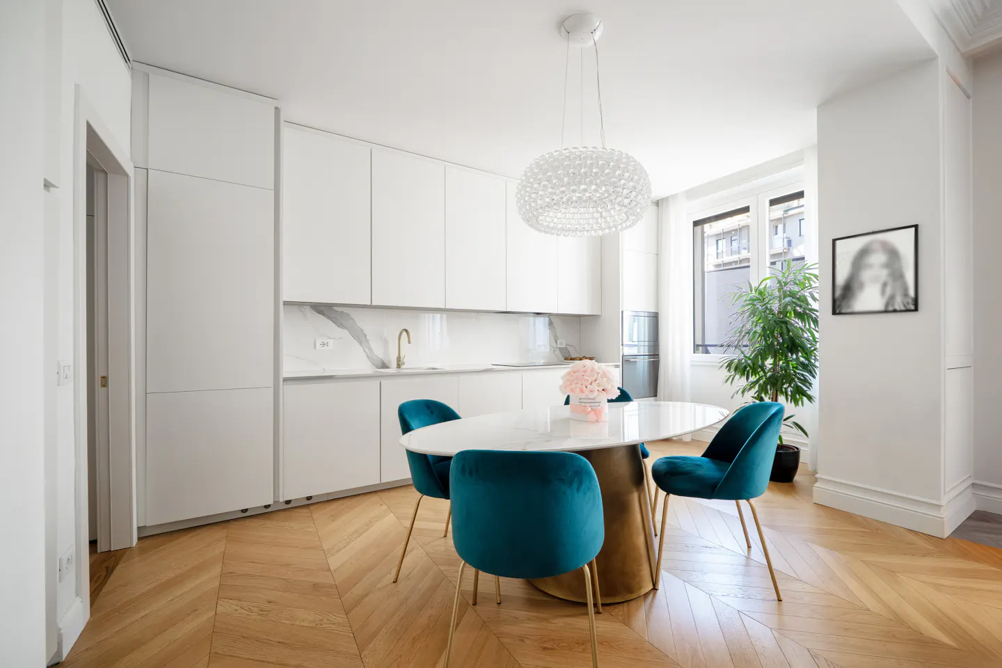 Bright, modern kitchen with white cabinets, marble counters, and herringbone wood floors. A round table with blue velvet chairs sits under a crystal chandelier.