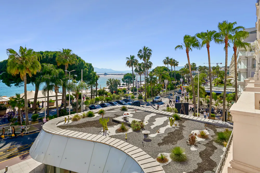 View from a balcony overlooking a coastal city street with palm trees, cars, and the ocean in the background under a clear blue sky.