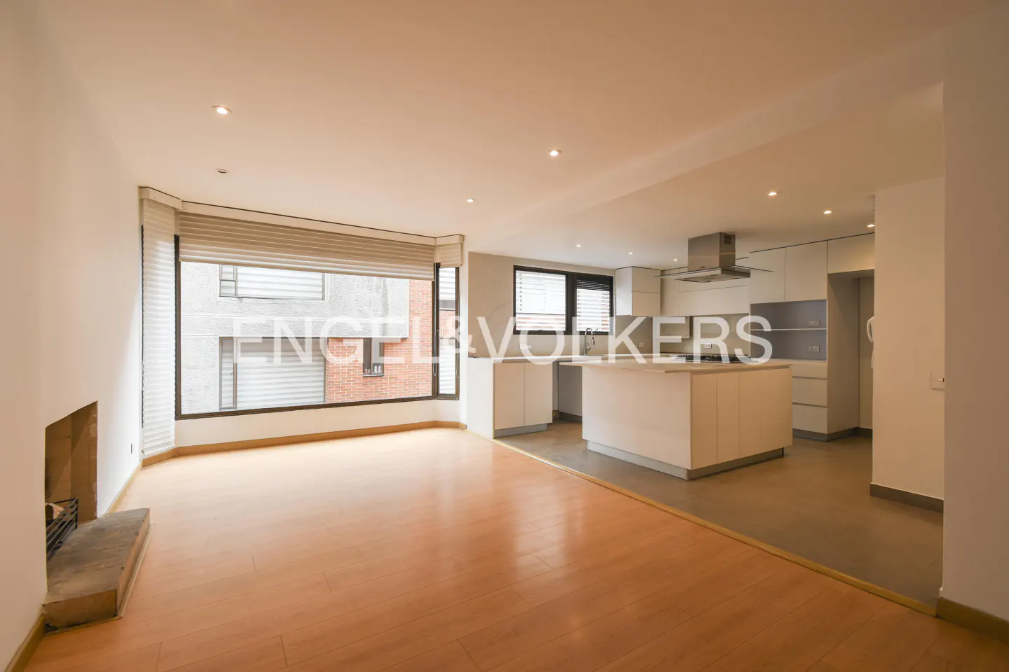 Open-concept living space with wood floors, a fireplace, and a modern white kitchen with an island. Large window with blinds.