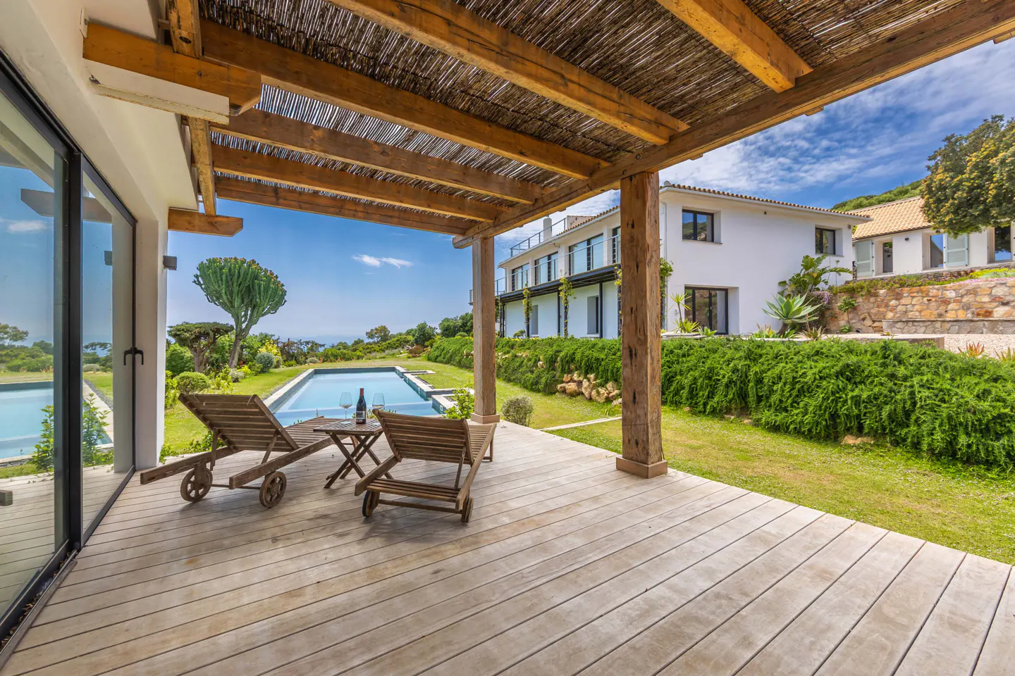 Wooden deck with lounge chairs, wine, and pool overlooking the ocean. White house and blue sky in the background.