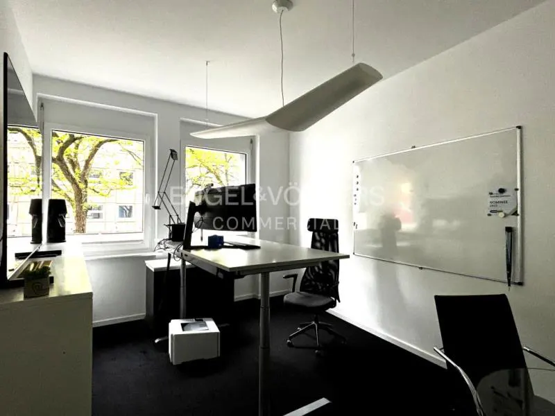 Bright office space with a desk, chair, whiteboard, and large windows with green trees outside. A modern, wing-shaped light fixture hangs from the ceiling.