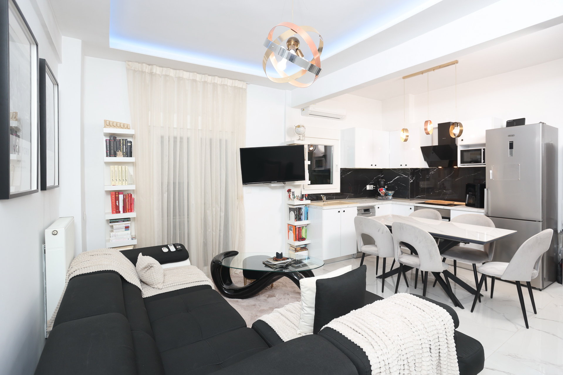 Bright, modern apartment interior with black sectional sofa, glass coffee table, and open kitchen with white cabinets and marble backsplash.