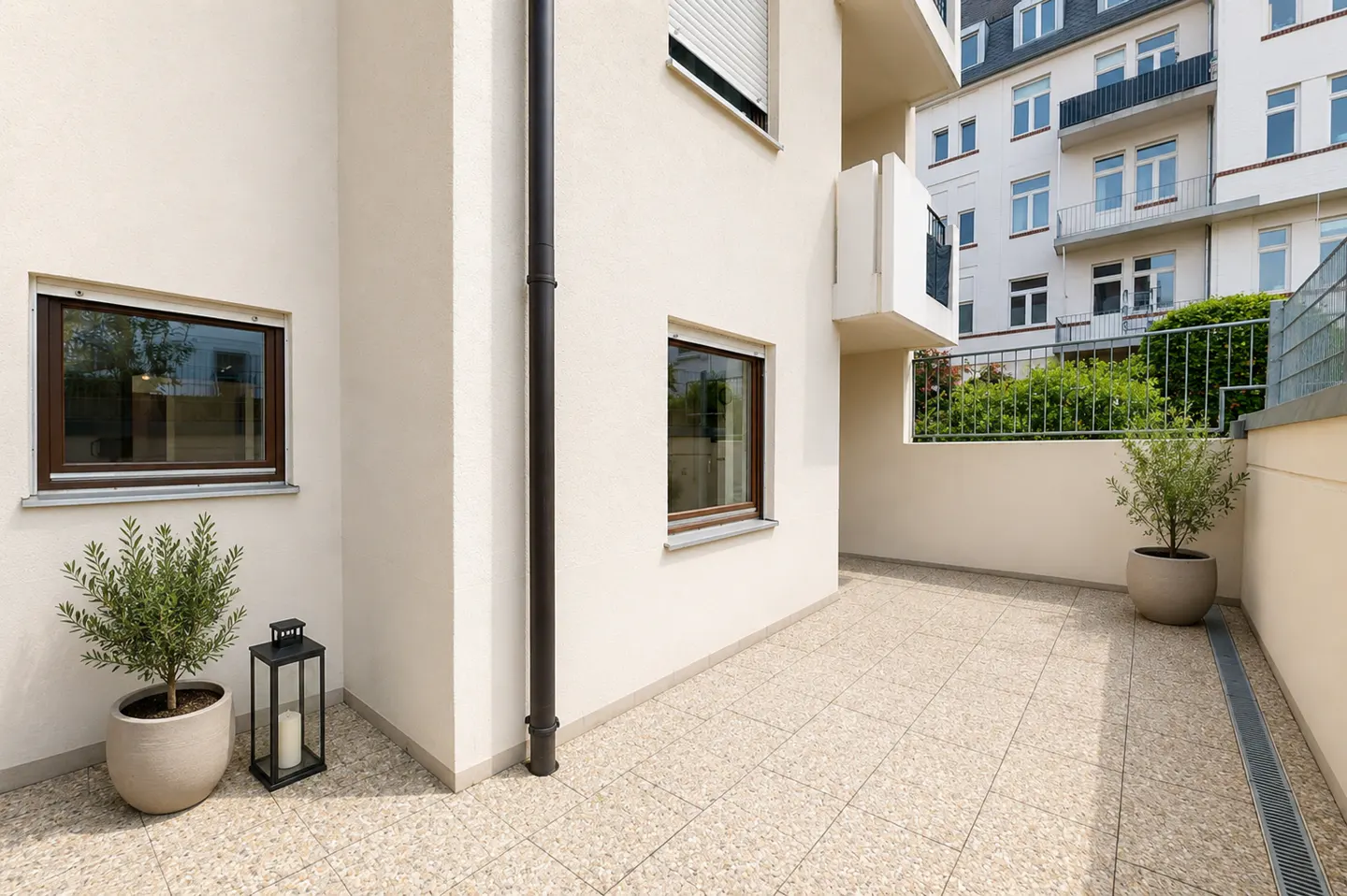 Exterior view of a beige building with a tiled patio, potted plants, and a black lantern. Windows with brown frames are visible.