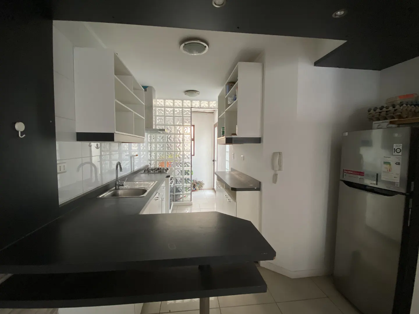 A bright kitchen with white cabinets, black countertops, and a stainless steel refrigerator. A glass block wall provides natural light.