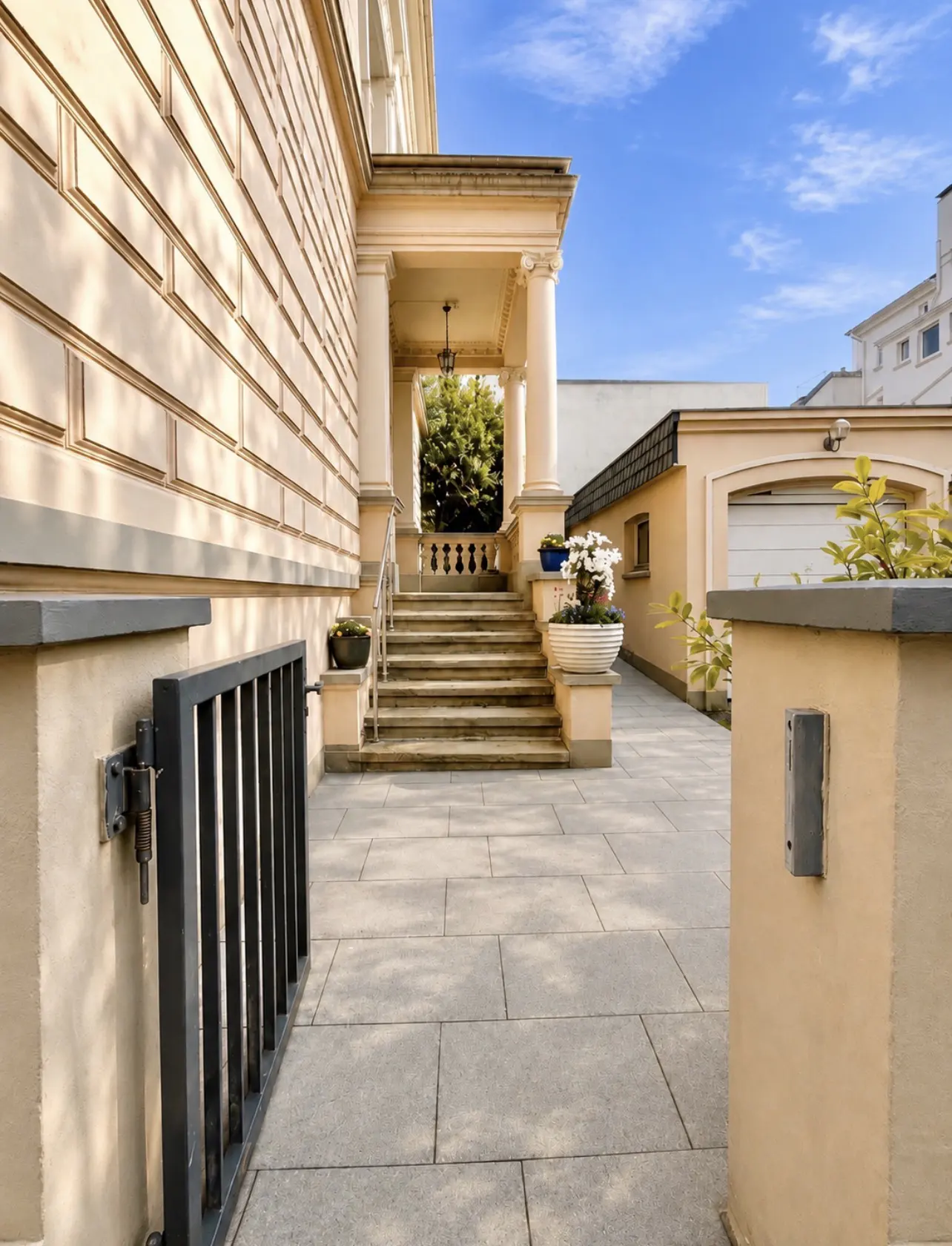 Exterior view of a beige house with a stone walkway leading to a columned porch and stairs. A black metal gate is on the left.