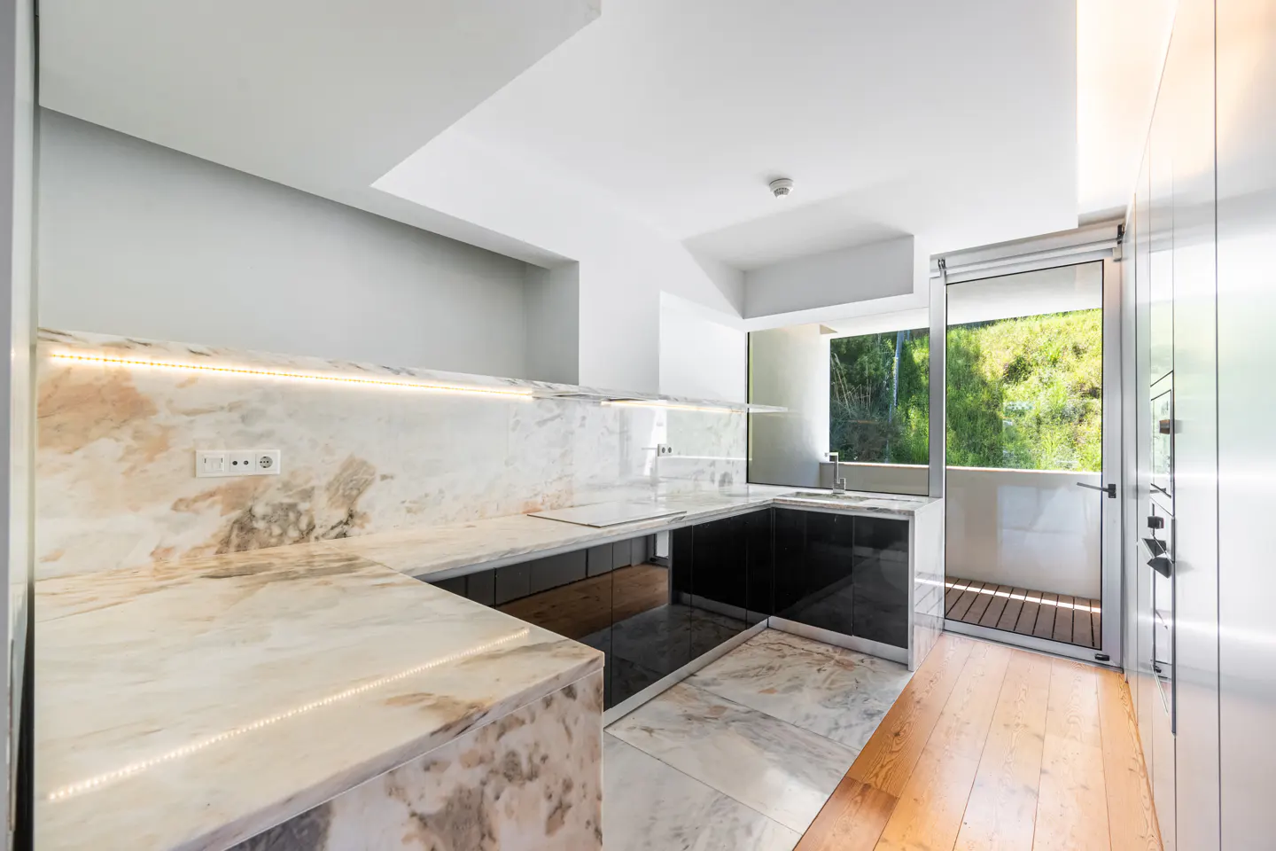 Bright kitchen with marble countertops, black cabinets, and wood floors. A glass door leads to a balcony with green trees in the background.