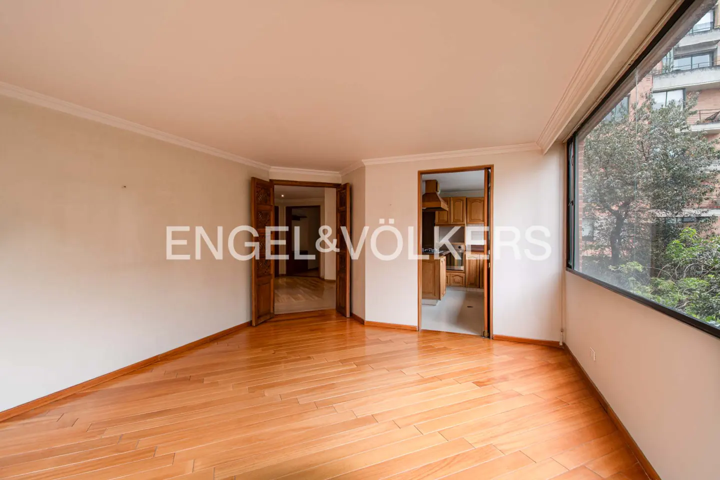 Bright, empty room with hardwood floors, large window, and two doorways leading to other rooms. Engel & Völkers logo visible.