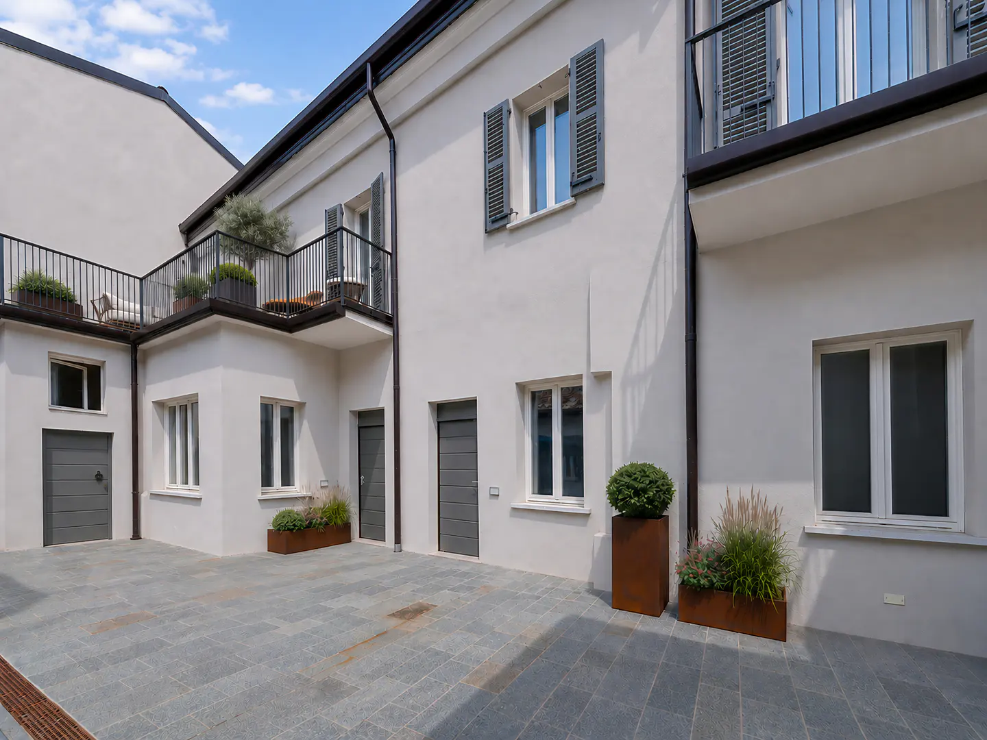Modern white building exterior with grey doors, windows with shutters, a balcony, and stone-paved courtyard with planters.