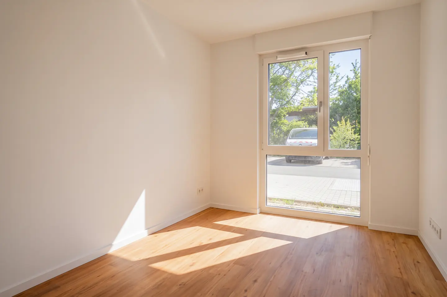 Bright, empty room with light wood floors and white walls. A window shows a car and trees outside. Sunlight streams in.