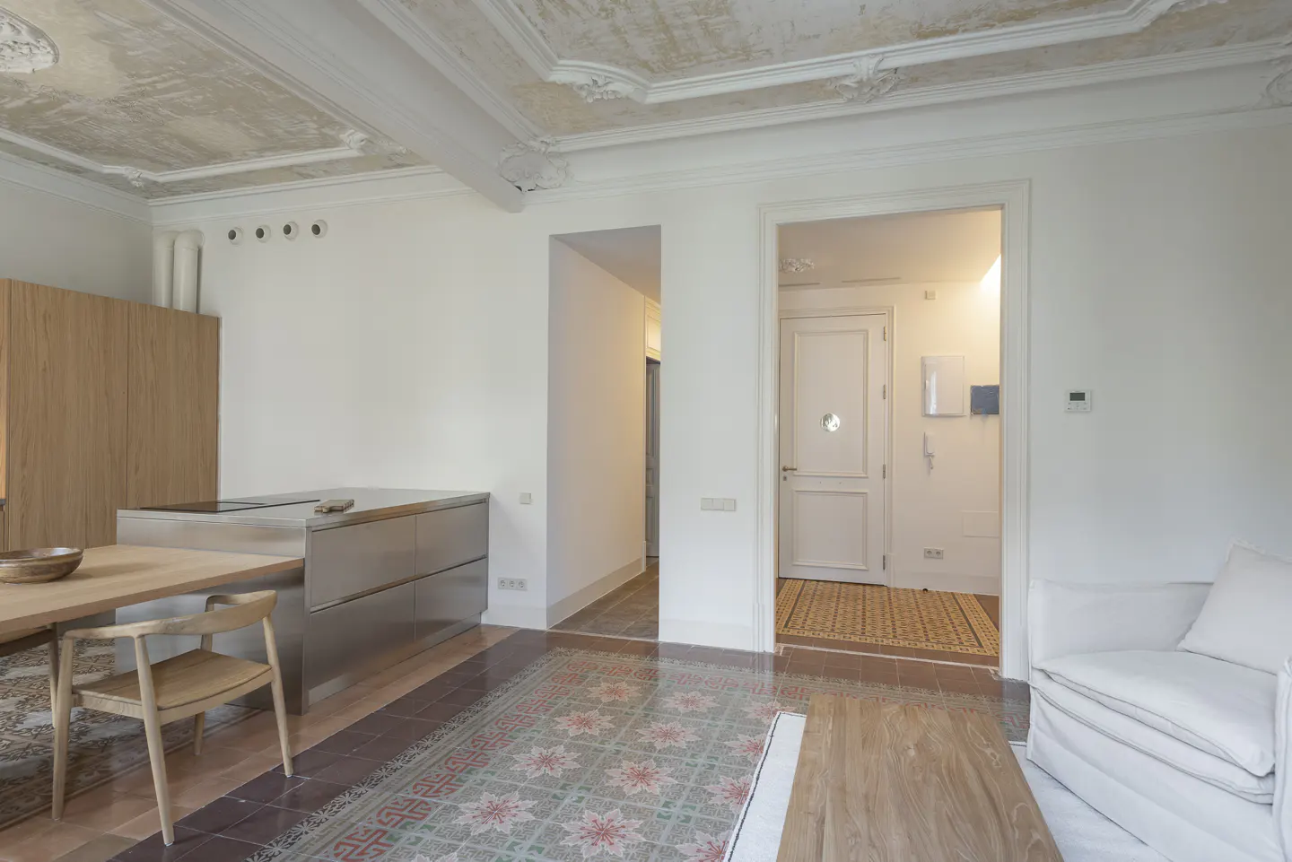 A bright, open-concept apartment with a kitchen, dining area, and living room. Ornate ceiling detail, colorful tile floor, and white walls.