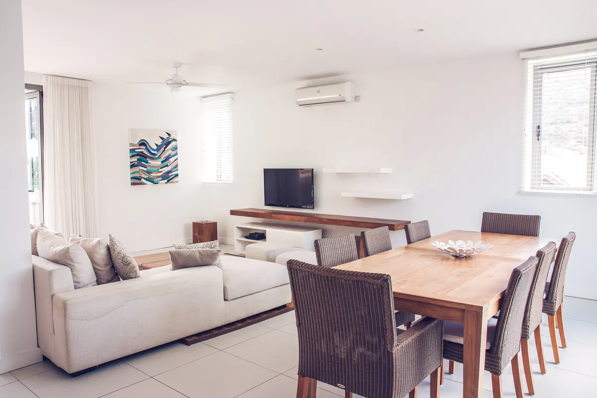 Bright living and dining area with white walls, tile floors, and a large sectional sofa. A wooden dining table with six chairs sits nearby.