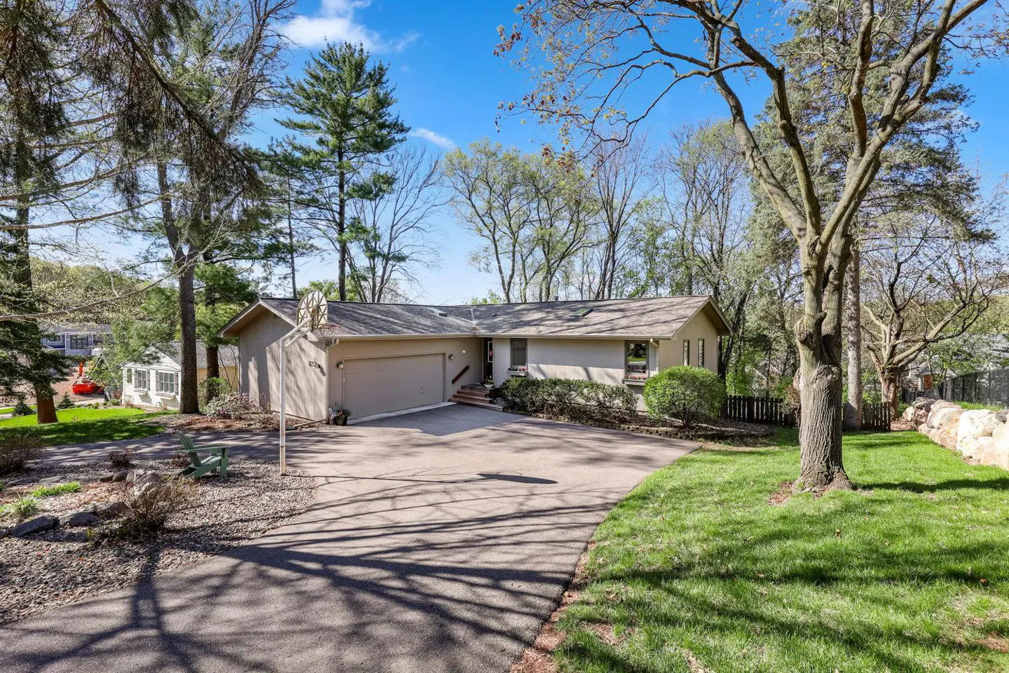 Beige ranch-style house with attached garage and asphalt driveway on a sunny day. Trees surround the property.