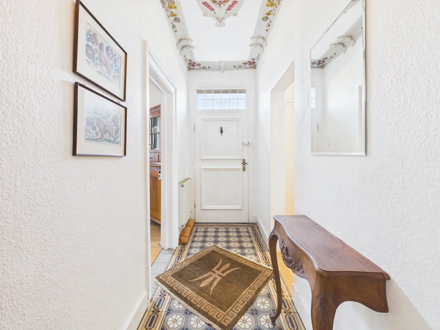 Hallway with white walls, tiled floor, and a brown console table. Two framed pictures hang on the left wall. A white door is at the end of the hall.