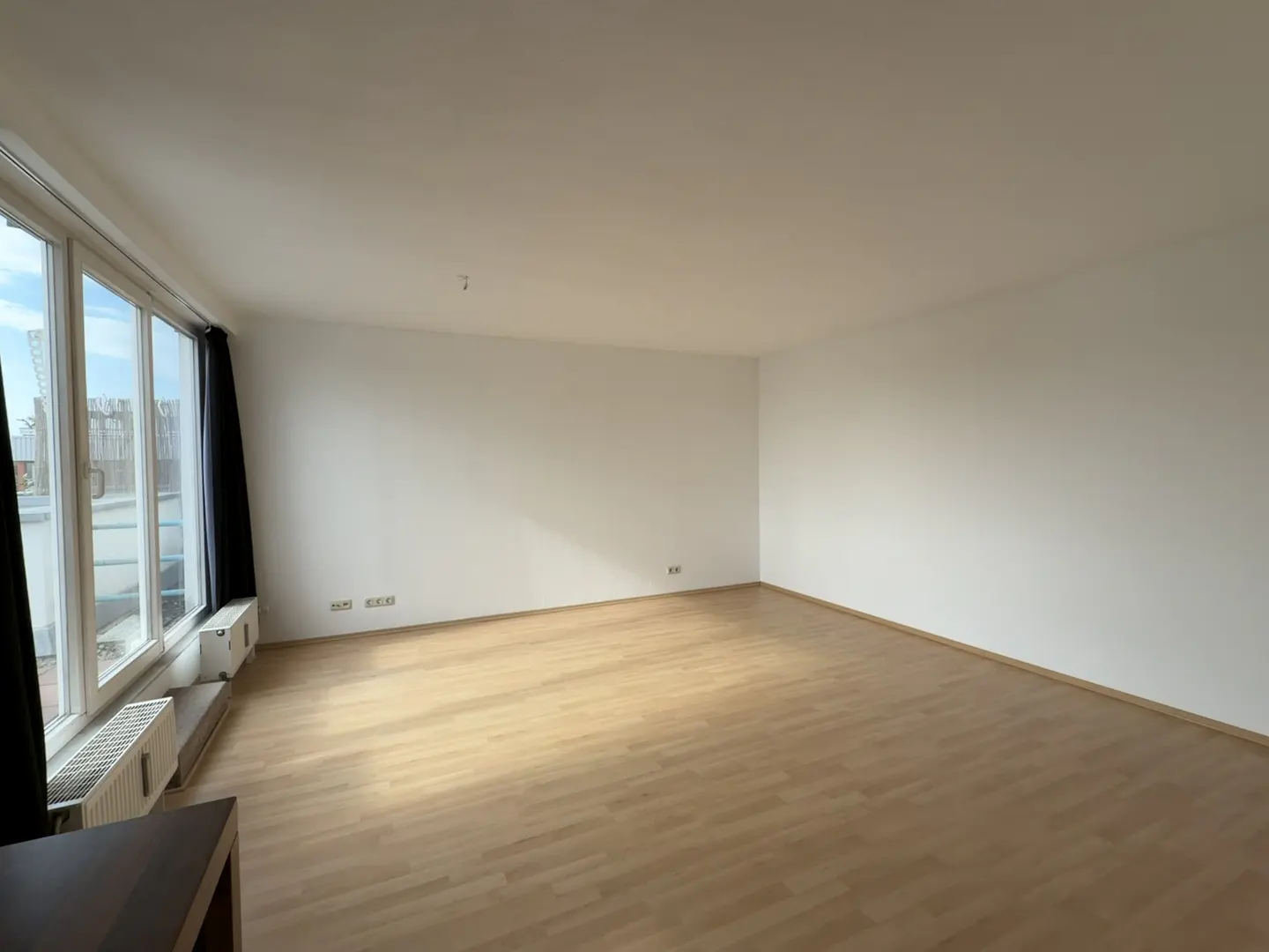 Bright, empty room with light wood floors, white walls, and large windows with black curtains. Radiators are visible under the windows.