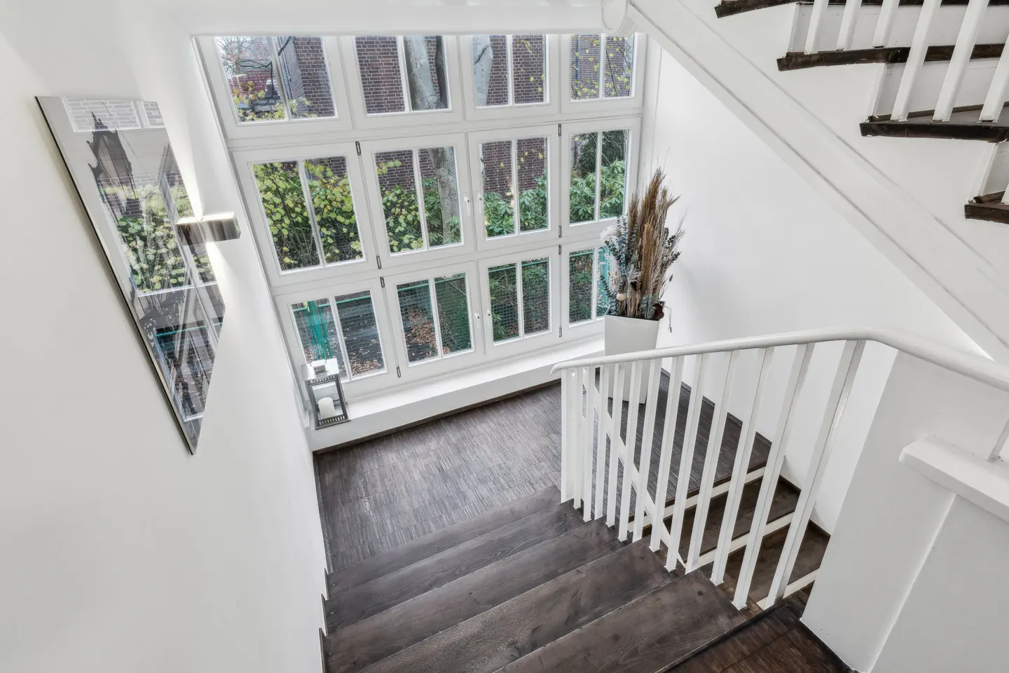 A high-angle shot of a staircase with white railings and dark wood steps, next to a large window and white walls.