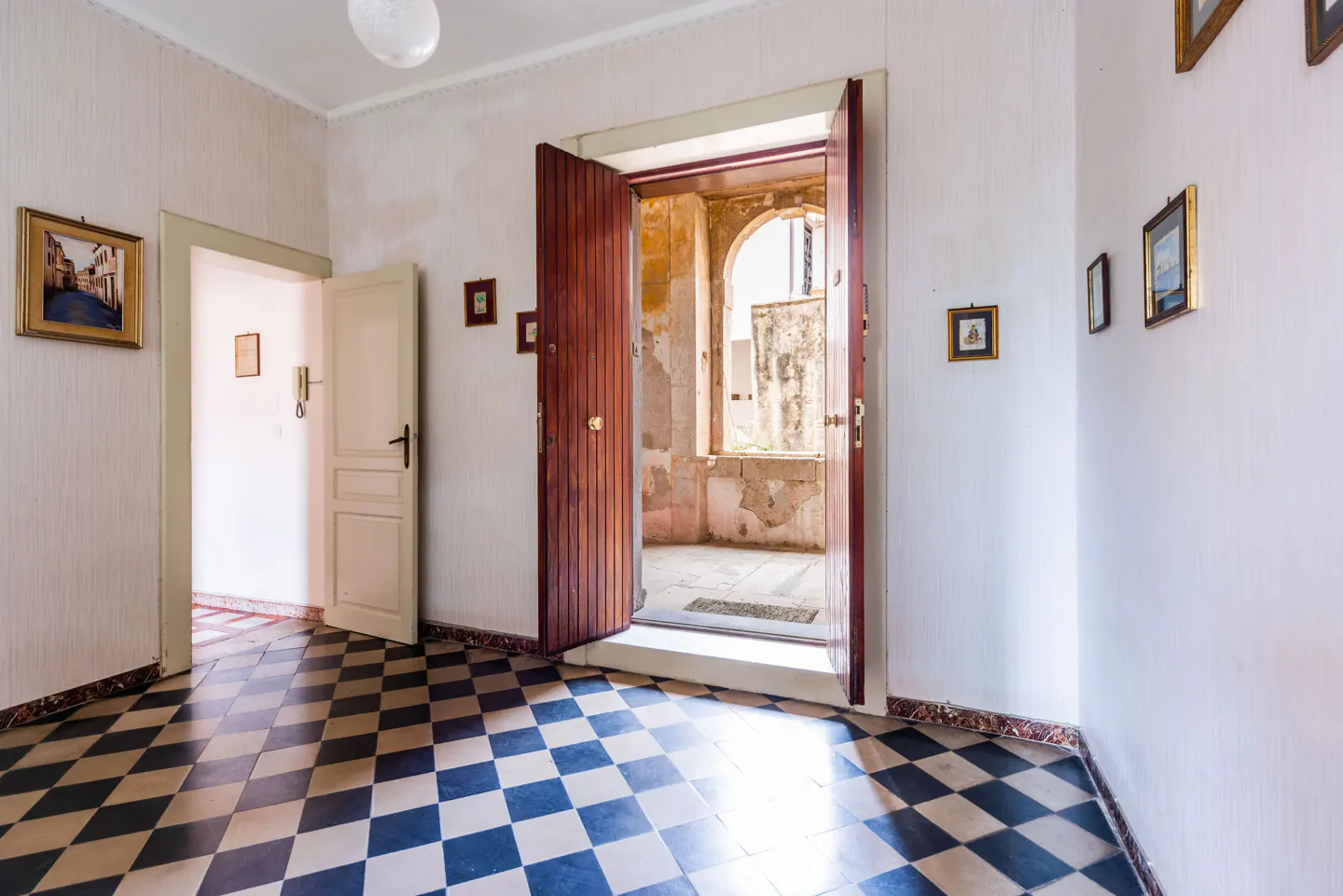 A bright hallway with black and white checkered floors, white walls with framed art, and an open wooden door leading to a sunlit room.