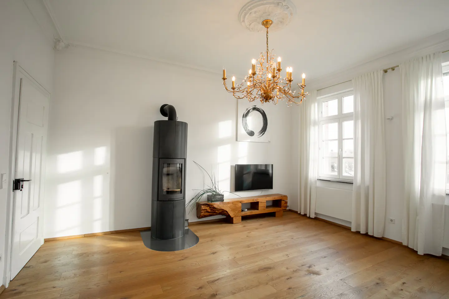 Bright living room with wood floors, white walls, and a black wood-burning stove. A gold chandelier hangs above a TV on a wooden stand.