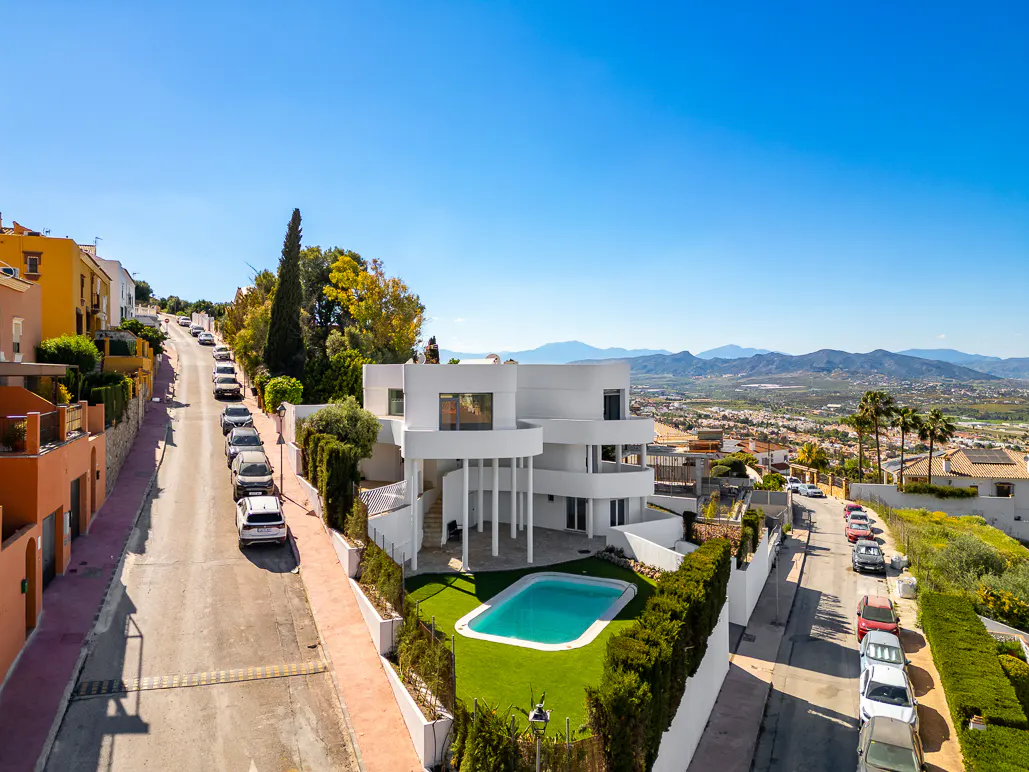 View of a modern white house with a pool on a hill, with a street and cars on either side. Blue sky.