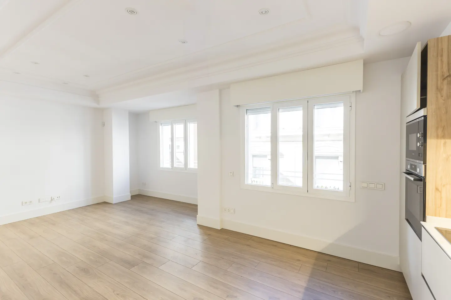 Bright, empty room with light wood floors, white walls, and two windows. A modern kitchen with built-in oven is visible on the right.