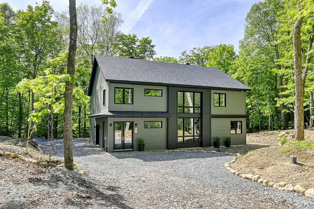 Two-story modern home with gray siding, black trim, and large windows, surrounded by lush green trees and a gravel driveway.