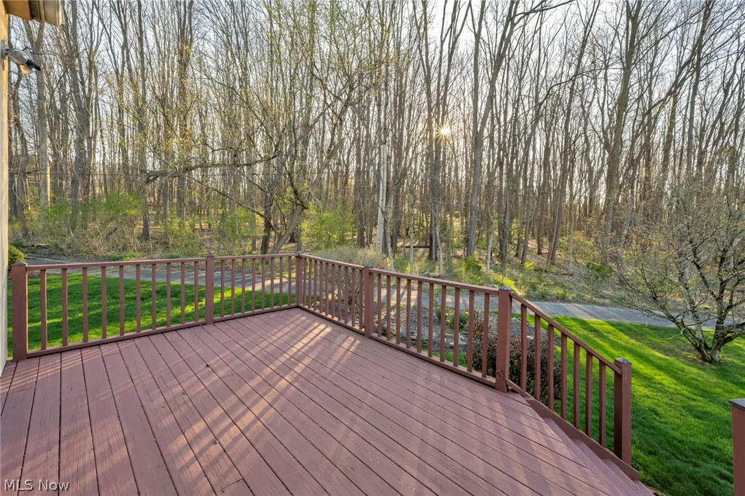 A brown wooden deck with railings overlooks a wooded area with green grass and a path. Sunlight filters through the trees.