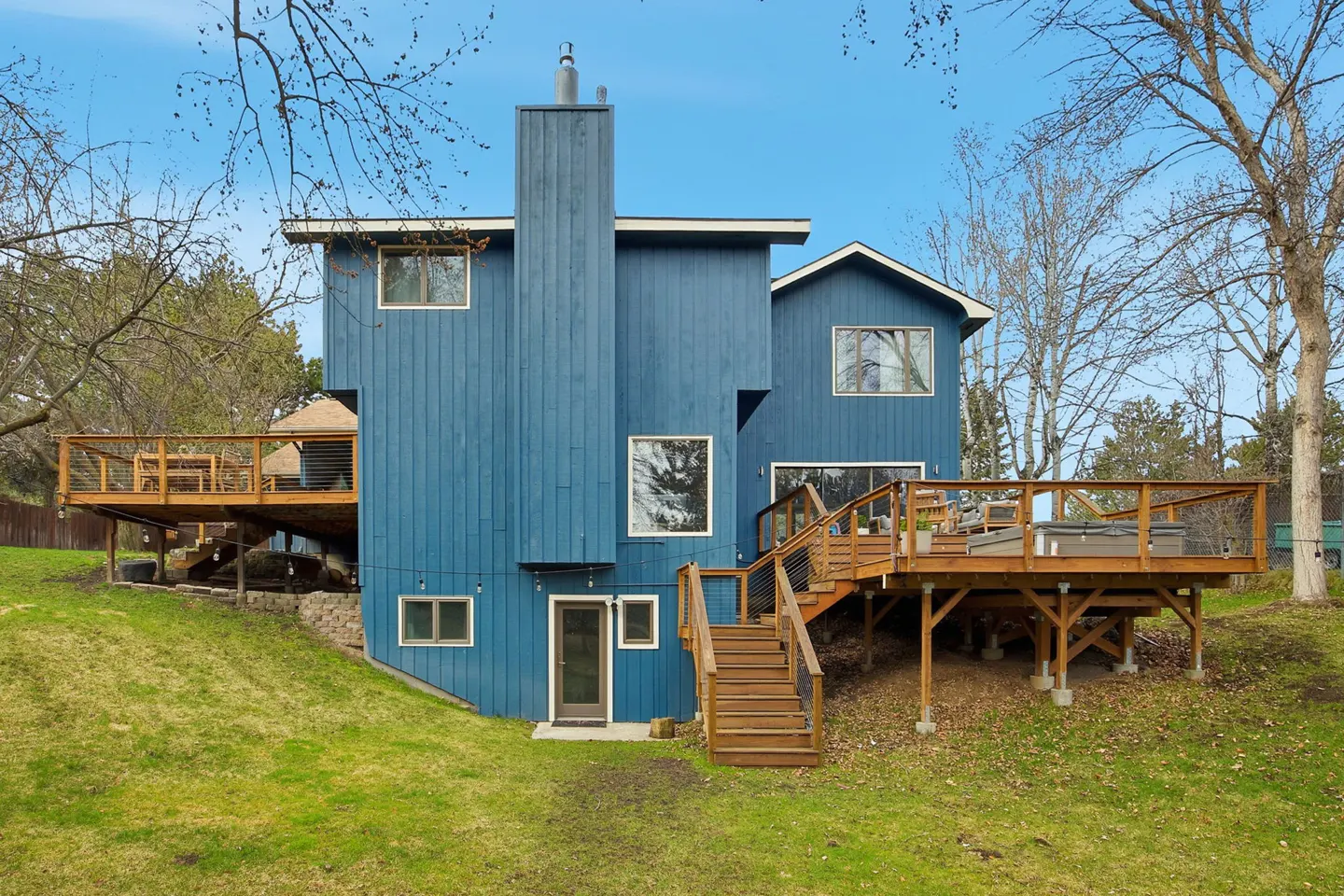 Back view of a blue two-story house with wooden decks and stairs on a green lawn.