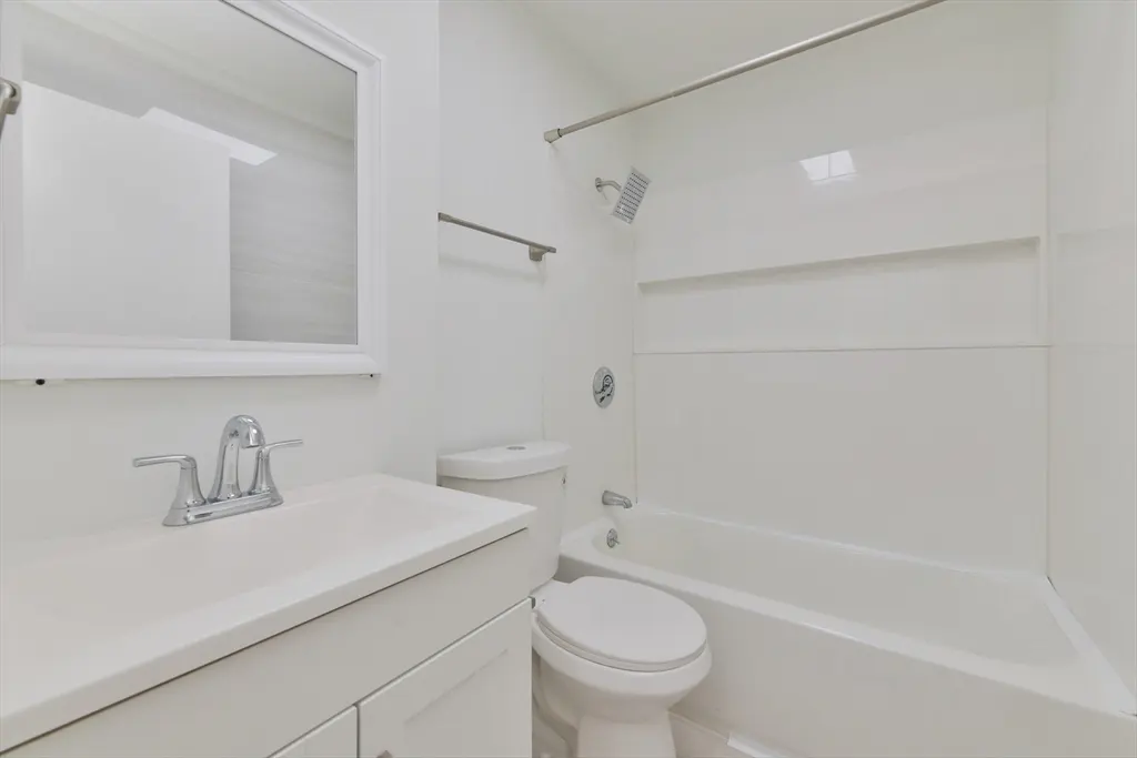Bright, all-white bathroom with a sink, toilet, and bathtub with shower. A mirror hangs above the sink.