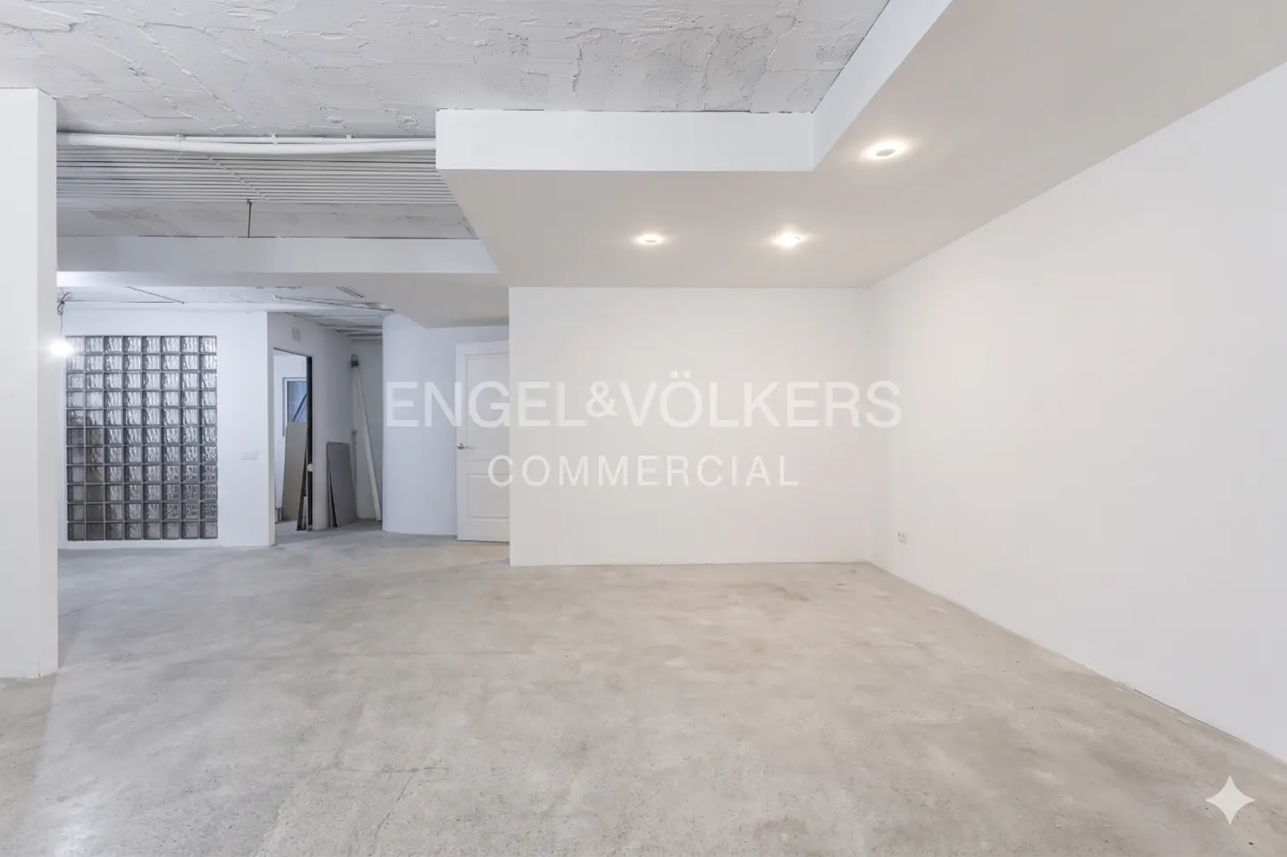 Bright, empty commercial space with white walls, concrete floors, and recessed lighting. Engel & Völkers logo is visible on the wall.