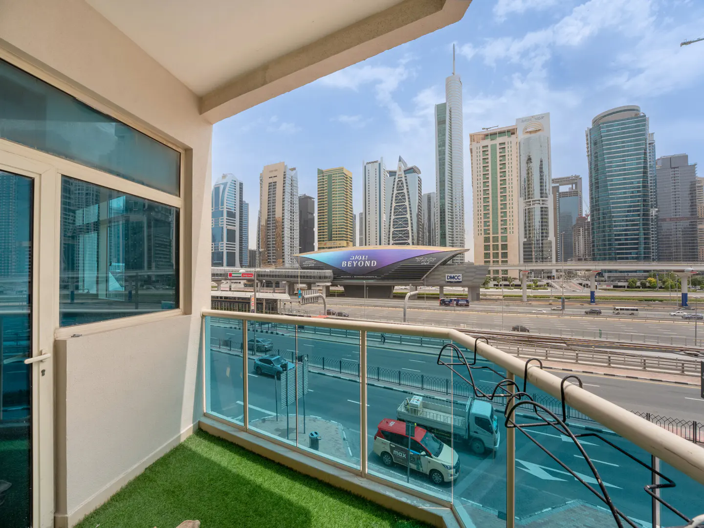 Balcony view of Dubai skyline. Blue glass railing overlooks a busy street with cars and a metro station. Green artificial turf on the balcony floor.