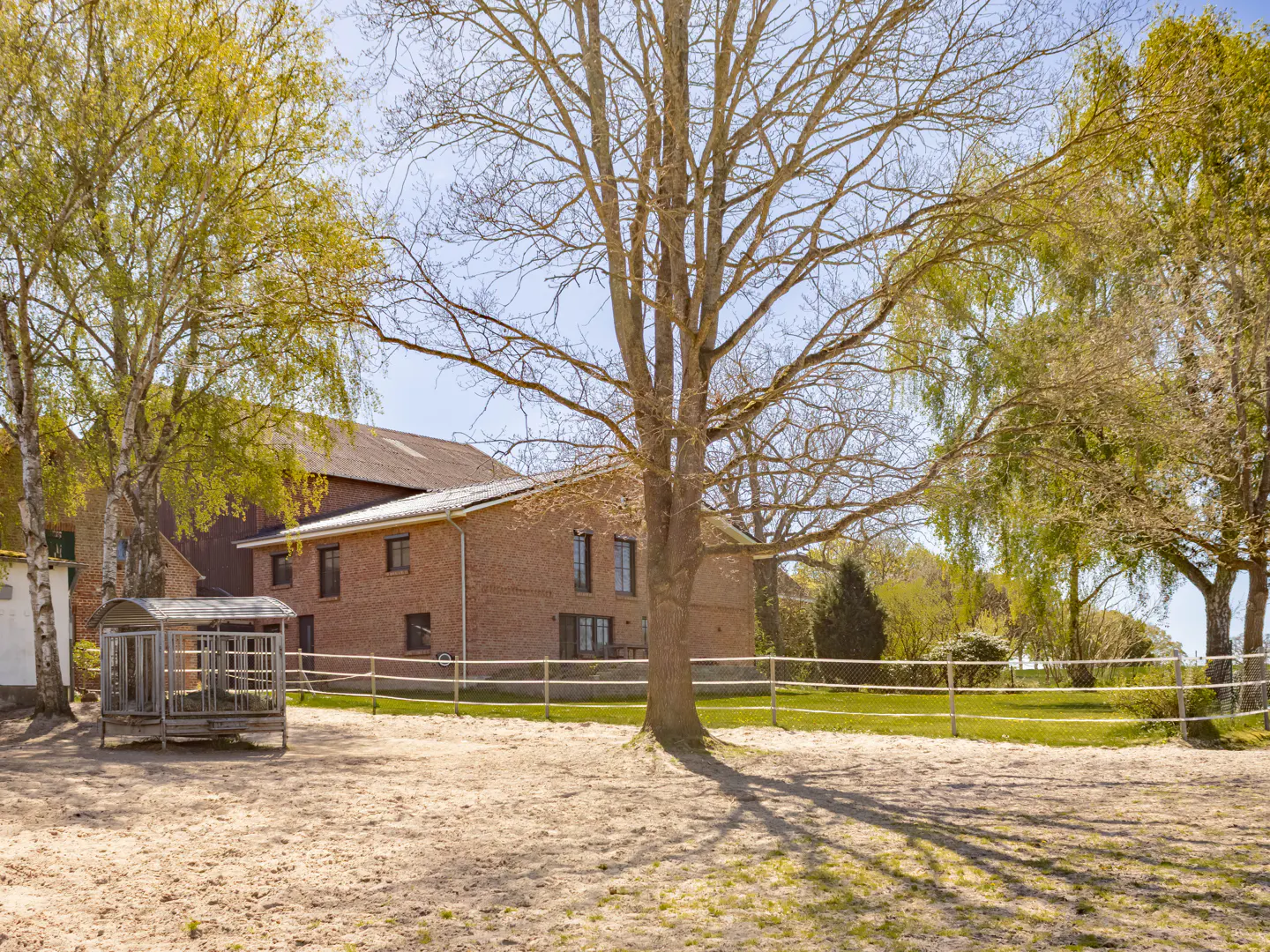 Exterior view of a red brick farmhouse with a sandy yard, trees, and a white fence on a sunny day.