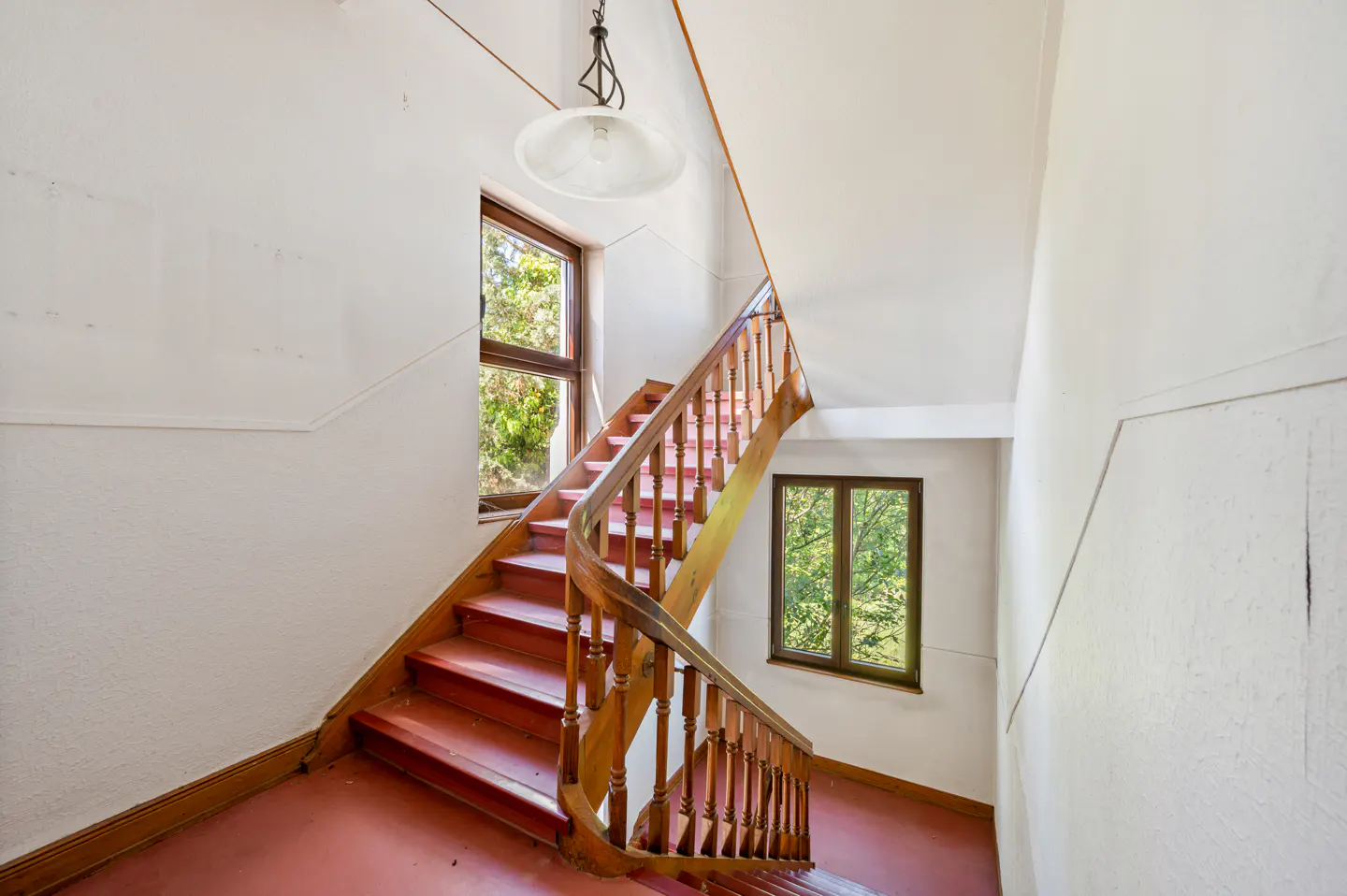 Interior view of a staircase with red carpet, wooden banister, and two windows with green trees visible outside.