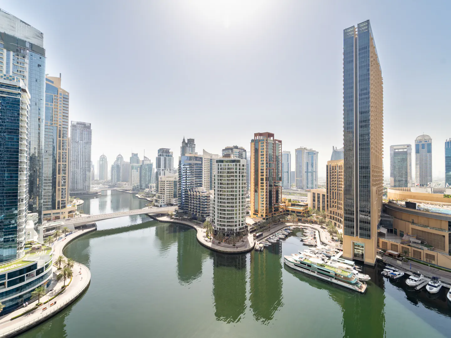 High-angle view of Dubai Marina with modern skyscrapers, a winding canal, and docked yachts under a bright, sunny sky.