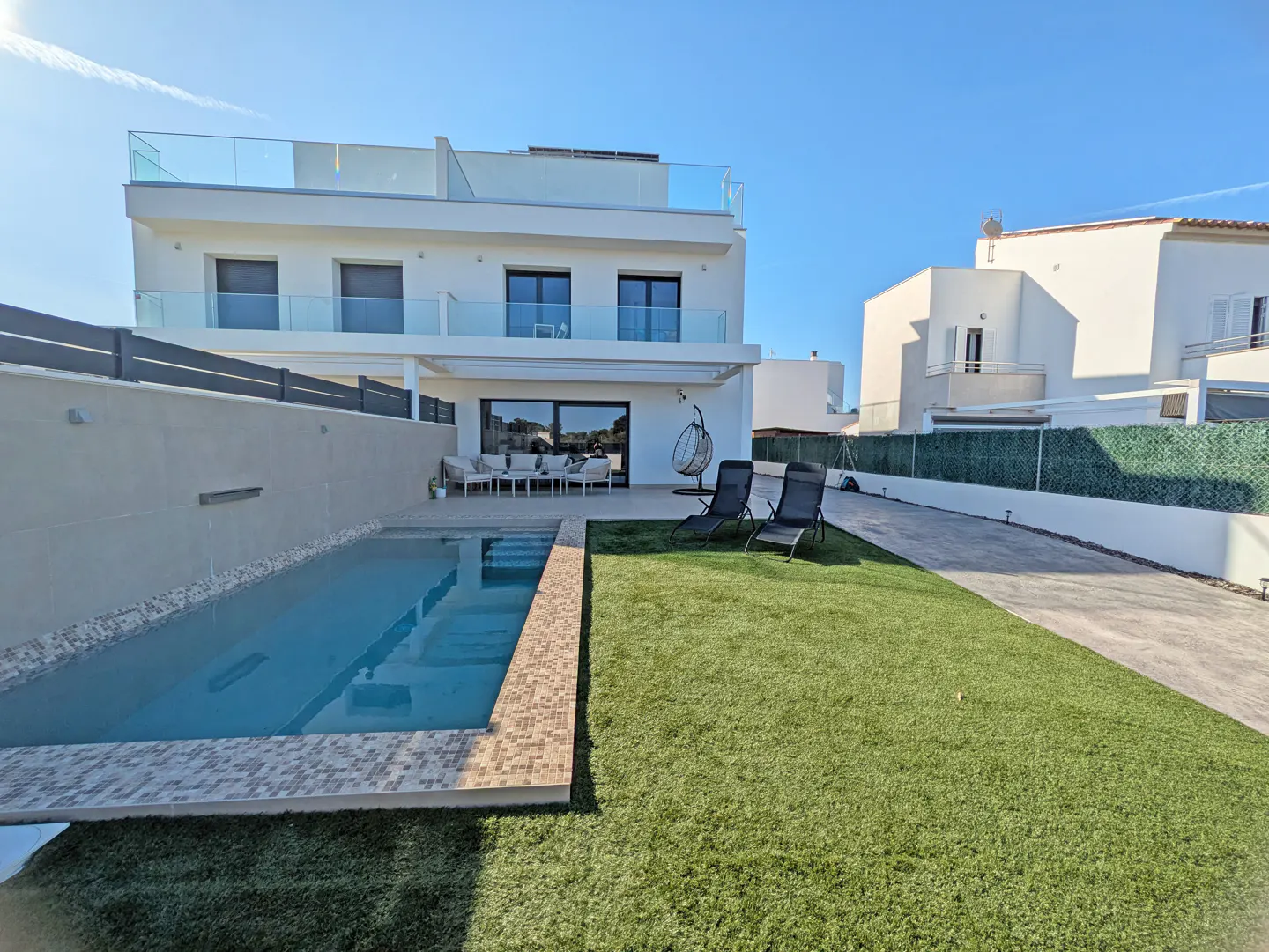 A modern, white two-story house with a pool and green lawn under a blue sky. Lounge chairs sit on the lawn.
